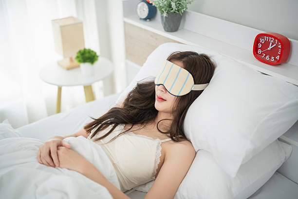 istock Young woman peacefully sleeping in bed with sleep mask, soft lighting, cozy white bedding, fresh green plants, and alarm clock on bedside table 2240045345