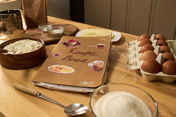 Recipe book surrounded by baking ingredients including flour in wooden bowl, eggs in carton, sugar in glass bowl, butter on plate, metal sifter and small glass bowls on wooden table