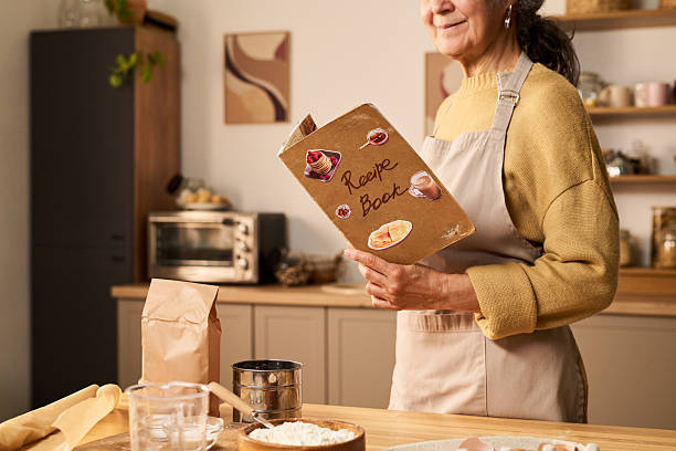 Middle aged Caucasian woman holding recipe book while preparing ingredients in kitchen, standing near counter with flour, sifter, paper bag and glassware, smiling slightly