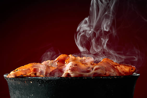 Fried steaming bacon slices in an old black pan on a dark red background. Copy space.