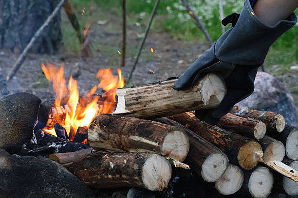 A horizontal photo of a hand in heavy welding gloves adding a log to a blazing campfire. Sparks rise into the air, capturing the warmth, energy, and authenticity of real outdoor camping life.