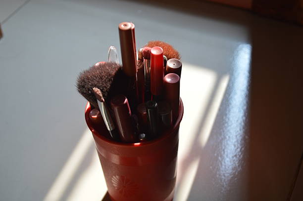 A collection of makeup brushes and cosmetic tools organized in a red holder on a sunlit vanity table. Soft light enhances colors and textures, highlighting beauty essentials for personal care and makeup artistry.