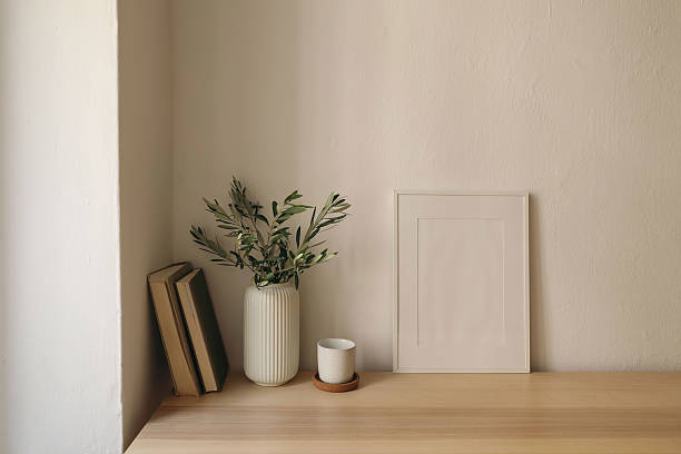 istock Vase with olive branches, old books. Cup of tea, coffee cork tray. Home office table. Blank white vertical picture frame mockup. Minimal Scandi Mediterranean interior. Beige wall background. 2240077506