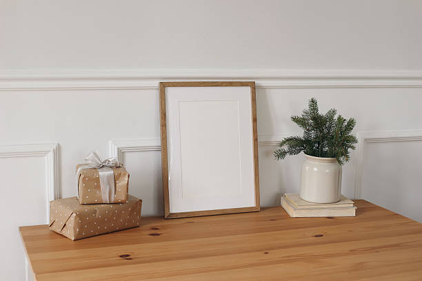 istock Christmas interior. Winter elegant artistic composition. Blank vertical wooden picture frame mockup. Gift boxes with ribbons on table. Fir spruce tree branches in vase, books. White wall background 2240077525