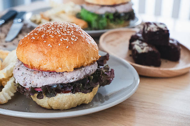 Vegetarian burger with sesame seed bun, fresh lettuce, and patty, served with crinkle-cut fries on a light grey plate, alongside blurred brownies on a wooden table.