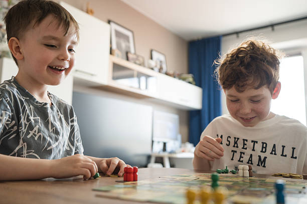 Two young friends share a fun moment playing a board game at home. Their joyful expressions underscore the positive interaction, teamwork, and the essence of leisure in a comfortable environment.