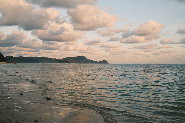 Ocean water gently lapping a sandy beach with distant mountains under a cloudy sky during a calm sunset
