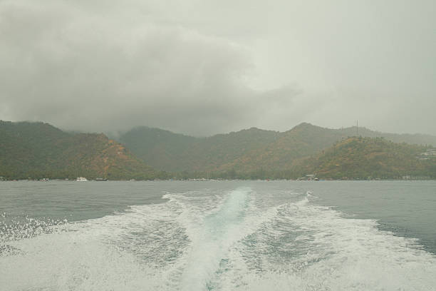 Boat wake trailing behind a vessel on the ocean, approaching distant tropical mountains under an overcast sky