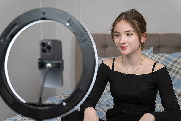 istock Young smile influencer sitting on bed, recording video content with smartphone and ring light, creating engaging material for her online audience 2240087760