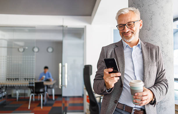 istock Mature businessman smiling while using smartphone and drinking coffee in office 2240093028