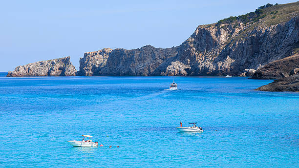 Horizontal landscape photo of bright blue sea with two to three boats floating on calm water. Majestic mountains rise on a distant island in the background under clear sunlight. Perfect for travel, nature, seascape, vacation, and coastal concepts.