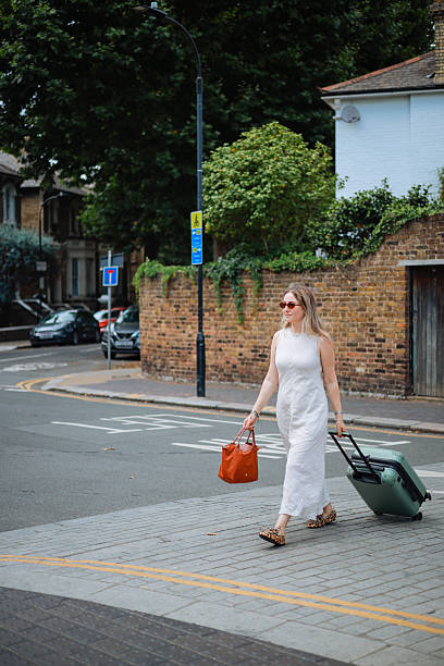 A happy adult black female entrepreneur on business travel in London. She is holding the handle of her large suitcase while standing on a sidewalk waiting for her taxi to take her to her hotel. She has an afro hairstyle and is wearing all black.