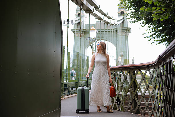 Woman walks confidently across a city bridge, pulling a green suitcase behind her. She is dressed in a floral top and white dress, enjoying her urban adventure with historic architecture and city life in the background, creating a vibrant travel scene.