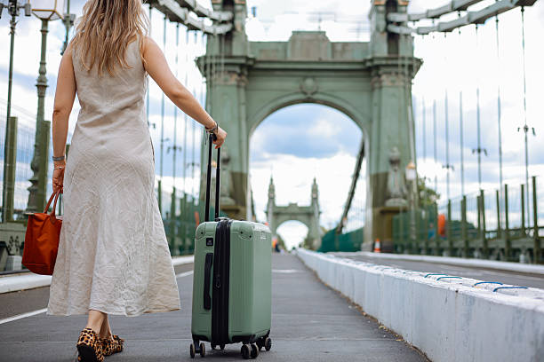 Woman walks confidently across a city bridge, pulling a green suitcase behind her. She is dressed in a floral top and white dress, enjoying her urban adventure with historic architecture and city life in the background, creating a vibrant travel scene.