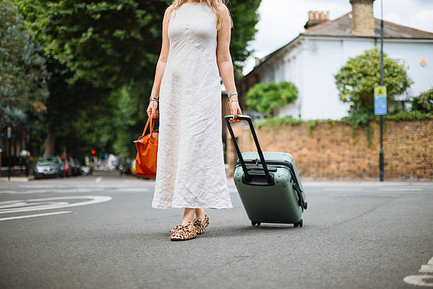 A happy adult black female entrepreneur on business travel in London. She is holding the handle of her large suitcase while standing on a sidewalk waiting for her taxi to take her to her hotel. She has an afro hairstyle and is wearing all black.