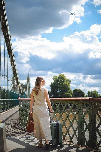 Woman walks confidently across a city bridge, pulling a green suitcase behind her. She is dressed in a floral top and white dress, enjoying her urban adventure with historic architecture and city life in the background, creating a vibrant travel scene.