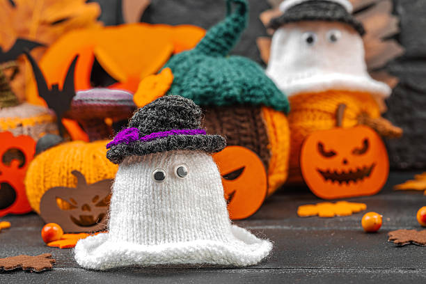 Cozy Halloween scene with cute ghost figurines in tiny hats. Handmade knitted autumn decor, crocheted pumpkins, fall leaves, berries, spiders, and ghosts. Dark brick background, place for text