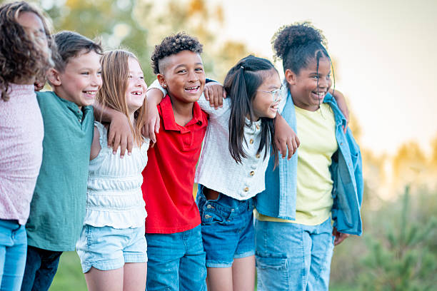 A cheerful, diverse group of children from different backgrounds links arms and smiles, capturing friendship, teamwork, and outdoor play in a sunlit park setting.