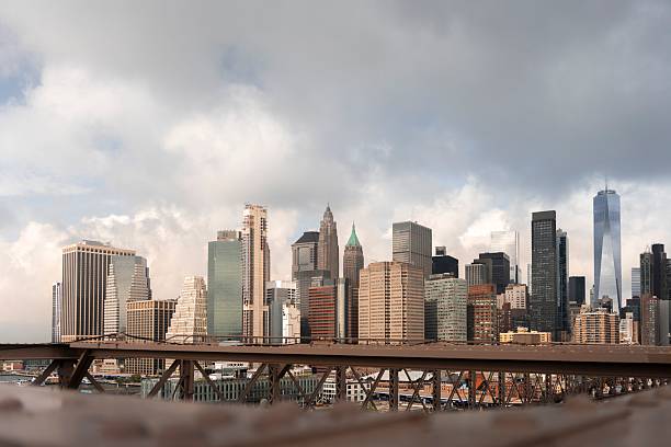View of the New York City skyline from the Brooklyn Bridge, featuring iconic skyscrapers under a cloudy sky.