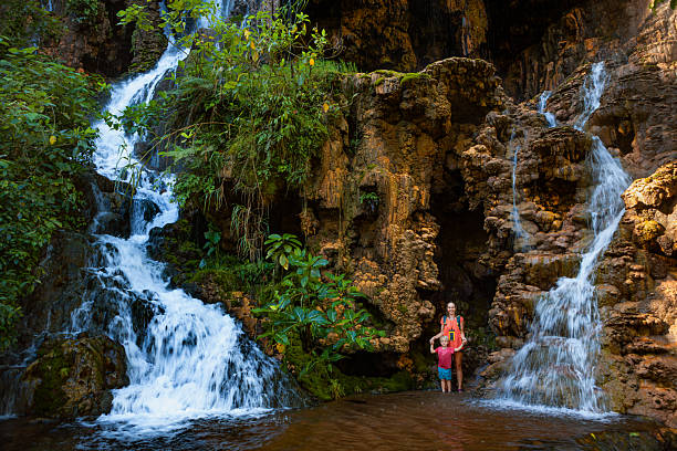 Mother with child standing under scenic Goa Tetes waterfall in East Java, Indonesia. Lush jungle surroundings, dramatic cliffs, and cascading streams create perfect backdrop for adventurous family travel experience in Southeast Asia.