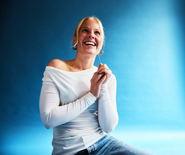 Smiling young woman with natural skin and blonde hair laughing brightly while seated in front of a blue background, radiating happiness and self-confidence.