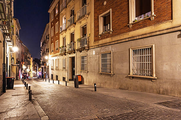 Madrid, Spain, Europe, September 13, 2025: A quiet side street in Madrid is captured on a warm autumn night, lined with multi-story residential buildings featuring balconies, barred windows, and mixed brick and stucco facades. The cobblestone pavement and bollards reflect traditional urban design, while soft street lighting casts a warm glow across the empty road. A few distant figures add subtle depth to the otherwise tranquil scene. Trash bins and signage hint at everyday residential life.
