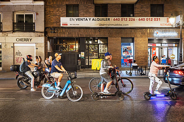 Madrid, Spain, Europe, September 14, 2025: A group of individuals on electric scooters, bicycles, skateboards, and motorcycles pause at a red light in a narrow Madrid side street during a warm autumn evening. The scene is illuminated by storefronts and streetlights, with signage and urban architecture framing the background. The variety of transport modes reflects the city’s dynamic blend of traditional and modern mobility.