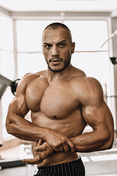 A shirtless, muscular man poses in a bright gym, showcasing defined arms and abs. Confident and focused, he projects strength, fitness, and athleticism ideal for sports or health-related campaigns.