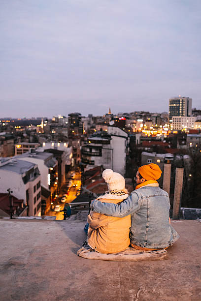 Rear view of a young couple enjoying view from a rooftop in winter clothing. Urban lifestyle scene with love, connection and city skyline in the background. City lights and copy space on the left