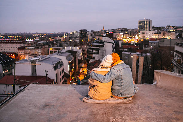 Rear view of a young couple enjoying view from a rooftop in winter clothing. Urban lifestyle scene with love, connection and city skyline in the background. City lights and copy space on the left