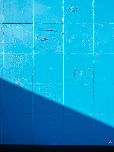 istock Blue tiled wall receiving diagonal shadow creating contrasting tones, showing textured surface with peeling paint, vibrant abstract background with bright sunlight 2240131091