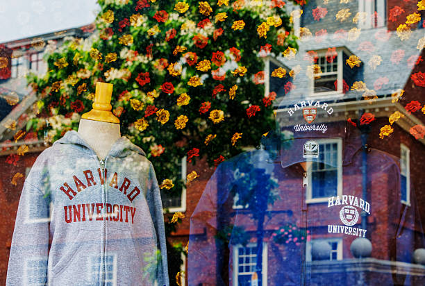 Cambridge, Massachusetts, USA  - June 8, 2025: Sidewalk view of a Harvard Square retail store window display of Harvard University brand clothing and souvenirs. Scene across the street reflected in the display window glass.