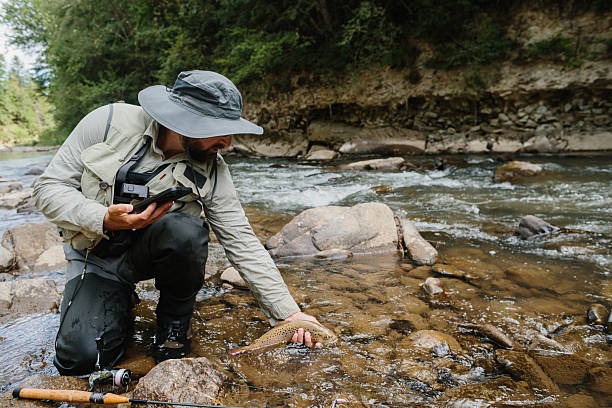 Fisherman holding a brown trout in his hand, about to release it back into a river, with fishing rod and phone in waterproof case visible