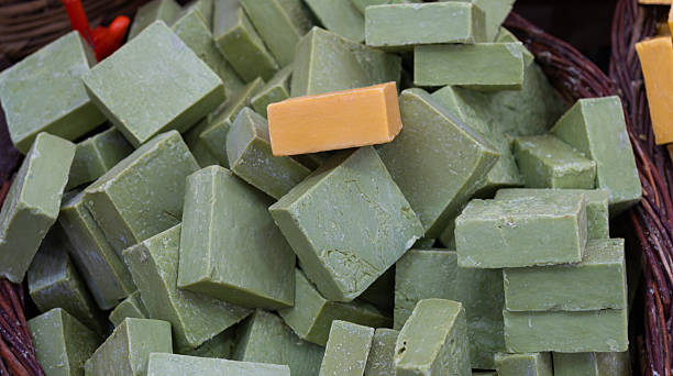 A rustic, close-up view captures several blocks of vibrant green soap, likely handcrafted or artisanal, neatly arranged within a traditional woven market basket. The soft, natural lighting highlights the soap's deep color and subtle texture, creating an inviting visual that strongly evokes themes of natural ingredients, local commerce, and traditional personal care products found at a bustling public market.