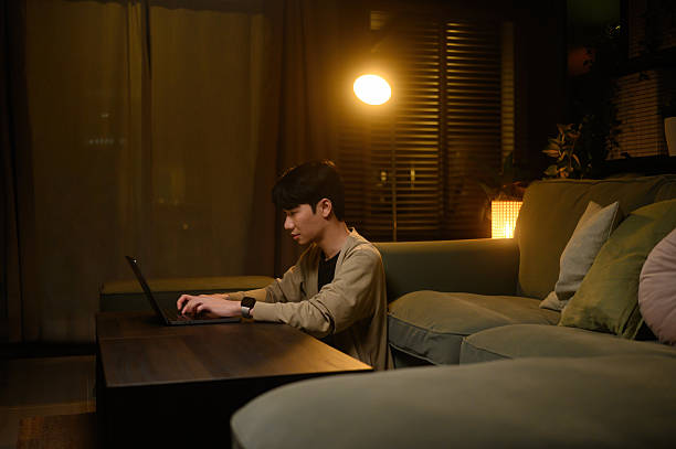 Asian man working on his laptop at home during nighttime, illuminated by warm lamp light, showing focus and productivity in a cozy living room