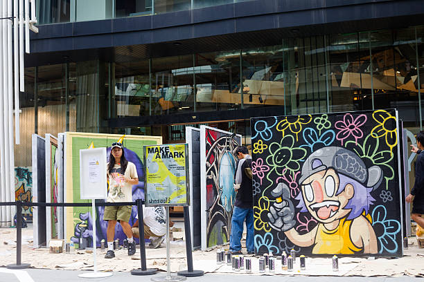 Group of thai graffiti artists is creating art in front of office building in Bangkok downtown. Scene is  at Phloen Chit Road nearBTS station. Artists are  creating artwotk in public over couple of weeks. One artist is standing behind a sign. He is holding a cup with iced drink and is looking at camera. Graffiti art is made on billboards. There is a an information sign about art creation and graffiti as freedom of language. Two other artists are spraying at different billboards