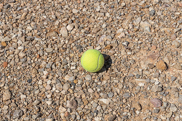 High angle view of a yellow tennis ball on a clay tennis court, with a shadow in front of it