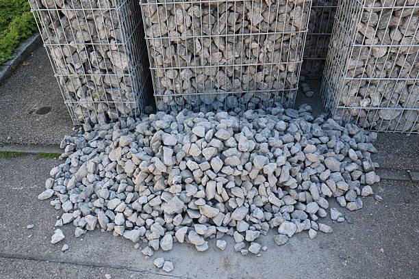A pile of rocks is on the ground next to a wall. The rocks are grey and there are three piles of them. .
