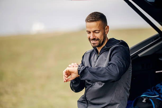 Smiling man in sportswear checking his smartwatch, standing near car trunk, preparing for or finishing an outdoor workout