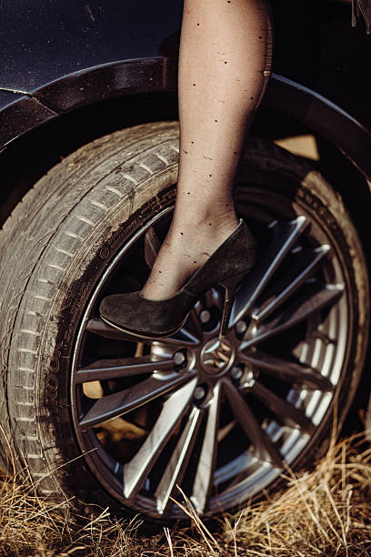 A woman's leg in black stockings, wearing black high-heeled shoes. The heel is elegant and slender. The shoe rests on the front tire of the car. The tire is black from dirt and wear. The wheel is silver. The woman's leg is partially visible, disappearing into the upper part of the frame. The body of the car is visible on the left, with a shiny, dark appearance. At the bottom of the image, dry, brown grass is visible, hinting at the outdoor setting. The lighting is natural, soft, slightly diffused. The color palette is mostly muted, with black, brown and silver tones. The perspective is low, focusing on the details of the shoe and the wheel. lifestyle image.