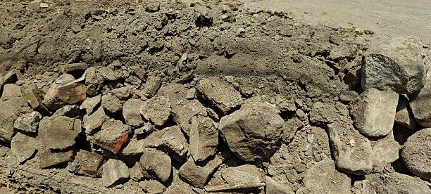 High-angle view of a deep, narrow construction trench or drainage ditch filled with loose soil and shadowed corners. The side wall shows layers of excavated earth and is topped by a large pile of concrete and brick rubble in harsh daylight