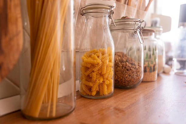 Close-up of various pasta types in transparent glass jars arranged on a wooden counter, highlighting textures and colors of uncooked spaghetti and fusilli in a warm kitchen setting.
