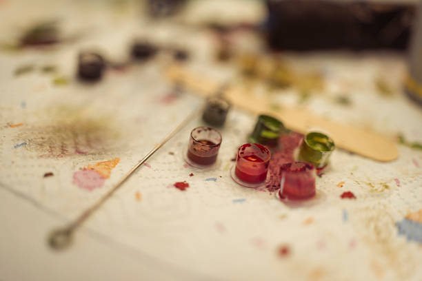 Close-up of vibrant tattoo ink cups in red, green, and other colors on a white surface, surrounded by scattered ink stains and tattooing tools in soft focus background.