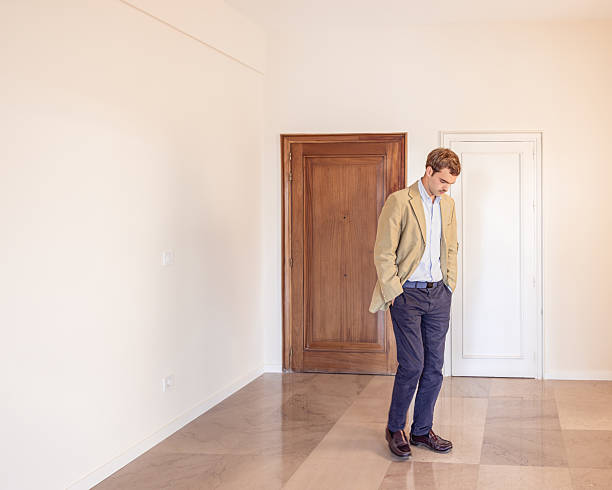 Man standing alone in an unfurnished room, hands in pockets, looking down