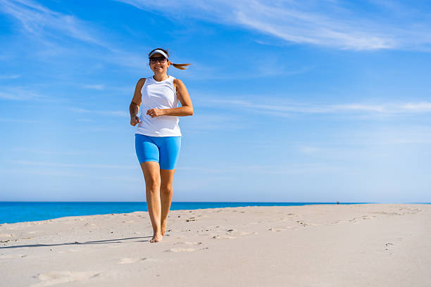 Physical activity on beach in summer. Mature woman running on sandy beach on sunny day. Front view