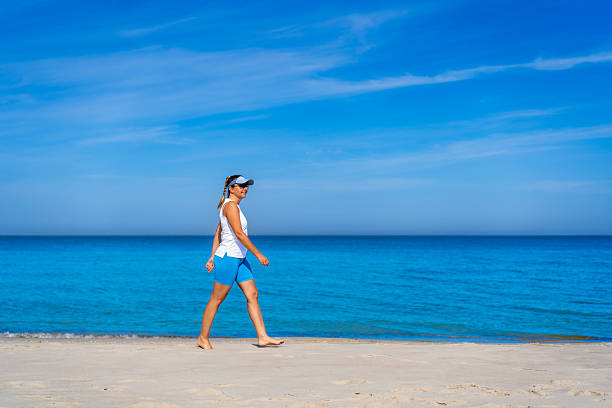 Physical activity on beach in summer. Mature woman walking  on sandy beach on sunny day. Side view