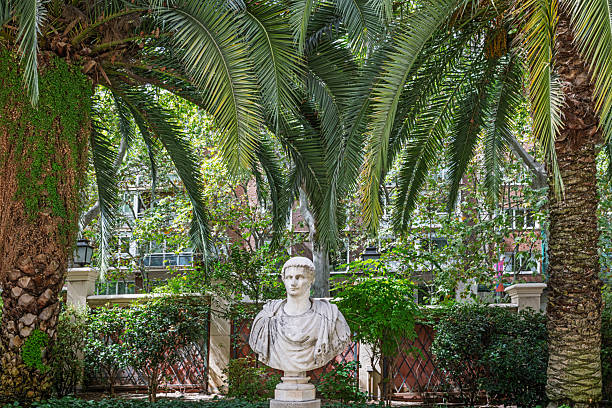 Madrid, Spain, Europe, September 13, 2025: Marble bust of a Roman-style figure displayed on a pedestal in a public park in Madrid, Spain. The sculpture is flanked by two tall palm trees and surrounded by lush greenery, with wrought iron fencing and architectural elements partially visible in the background. Natural sunlight filters through the foliage, highlighting the contrast between classical European art and Mediterranean vegetation.