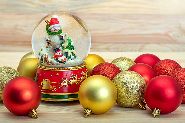 Large and small snowmen in a glass ball with snowflakes and red and gold Christmas decorations on a light wooden surface
