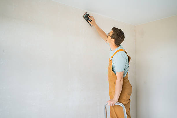 istock Construction worker wearing worn brown overalls, sanding white wall with electric tool while balancing on metal ladder during residential renovation work 2240231660