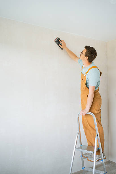 istock Construction worker in beige overalls balancing on metal ladder, sanding white wall with hand sander, preparing surface for smooth painting and renovation 2240232006